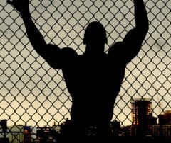 Outline of muscular man leaning against a chain link fence looking over a city