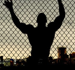 Outline of muscular man leaning against a chain link fence looking over a city