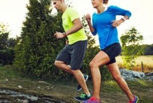 Couple wearing yellow and blue running uphill on dirt trail by meadow and bushes