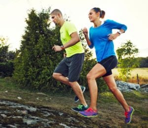 Couple wearing yellow and blue running uphill on dirt trail by meadow and bushes