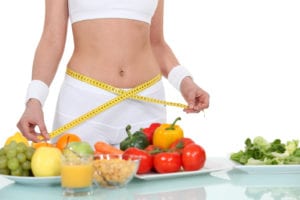 Woman in white workout clothes measuring hips with yellow tape measure by clear table with three plates of fruits, vegetables, orange juice, and cereal