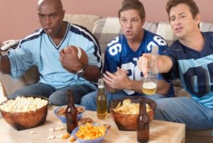 Group of three men in blue jerseys watching football on a couch with beers and snacks