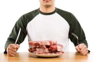 Man wearing green and white baseball shirt holding fork and knife sitting front of plate of four raw steaks