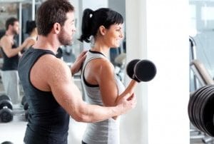 Young dark-haired young woman lifting dumbbell with help of young brown hair male personal trainer