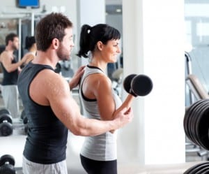 Young dark-haired young woman lifting dumbbell with help of young brown hair male personal trainer