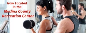 Black-haired young woman lifting weights with young male personal trainer helping in gym with red lettering about Medina County Recreation Center