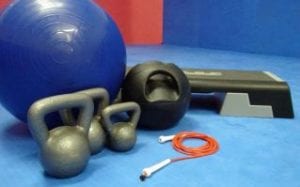 Three gray kettle balls, orange jump rope, blue exercise ball, black medicine ball, black step in blue and red gym