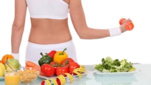 Woman in white workout clothes in lifting dumbbell behind table with veggies, fruit, cereal, orange juice, and yellow tape measure