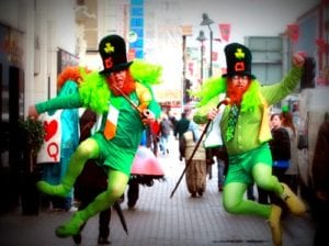 Two men dressed as red-bearded leprechauns in green doing heel click jump on city street