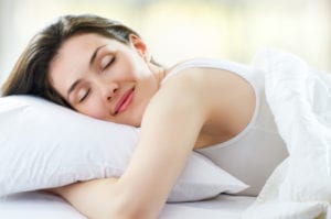 Burnette, smiling, young woman wearing white tank top hugging white pillow under white blanket sleeping in bed