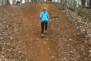 Young blonde woman walking up dirt trail with two dogs in the woods during late fall