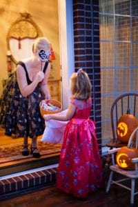 woman in black dress holding cany bowl out to girl trick or treating in pink dress