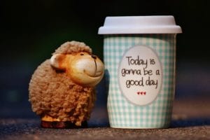 Small brown ceramic sheep by blue and white gingham printed travel coffee cup with inspiring quote on sidewalk