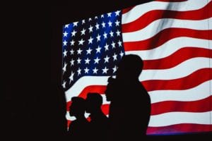 Shadow of four men standing in front of waving American flag on screen in black room