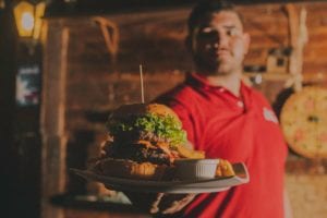 Waiter in red polo shirt holding close up of large burger and sauce in log cabin
