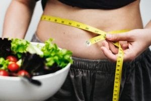 Person wearing gray and black baring belly to measure with yellow tape measure holding salad in white bowl