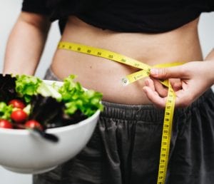 Person wearing gray and black baring belly to measure with yellow tape measure holding salad in white bowl