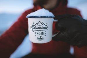 Person holding white and blue mug with with graphic and inspritional adventure quote filled with snow