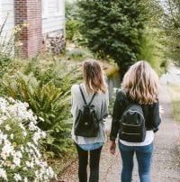 Two young women in jeans, sweater, and backpacks walking along garden near house