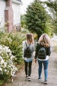 Two young women in jeans, sweater, and backpacks walking along garden near house